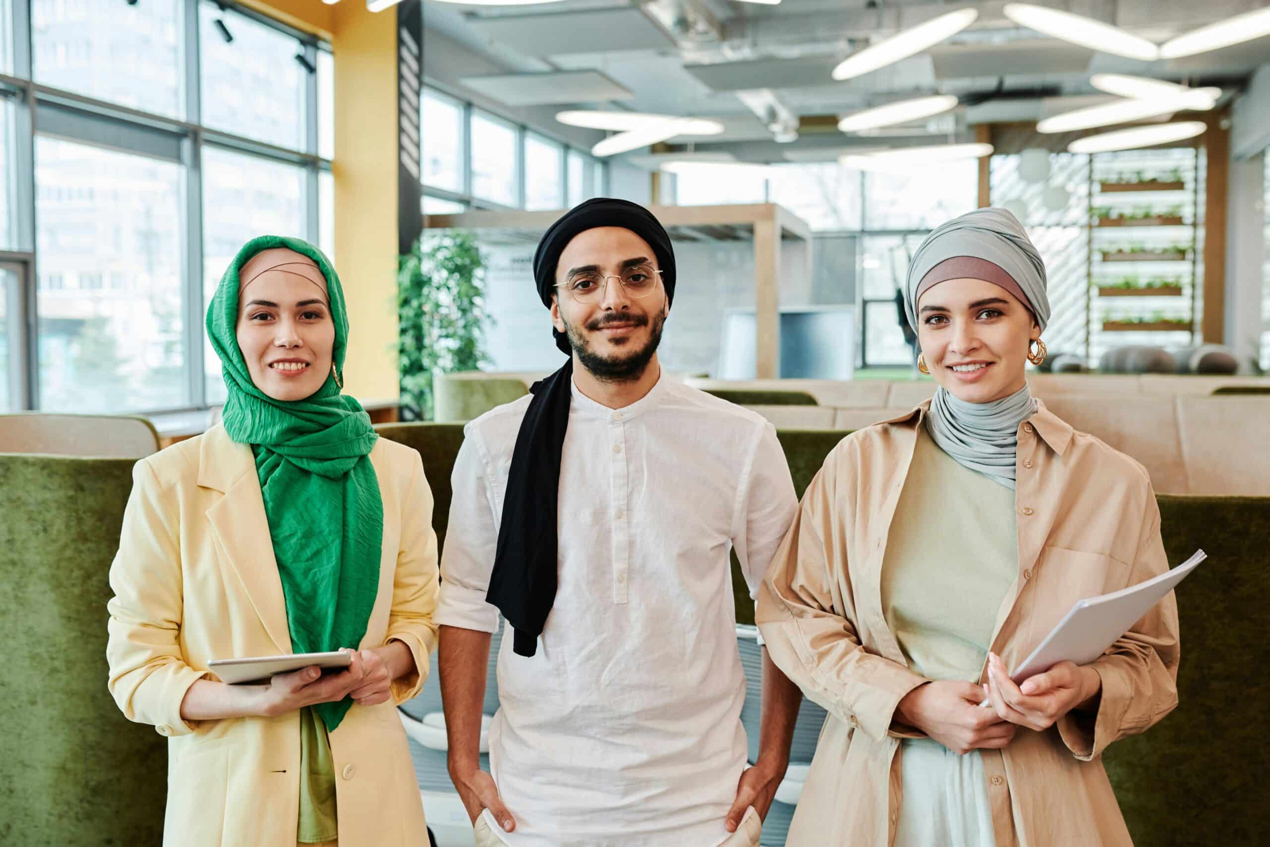 A diverse group of three professionals smiling in a modern office setting, wearing cultural attire such as headscarves and turbans.