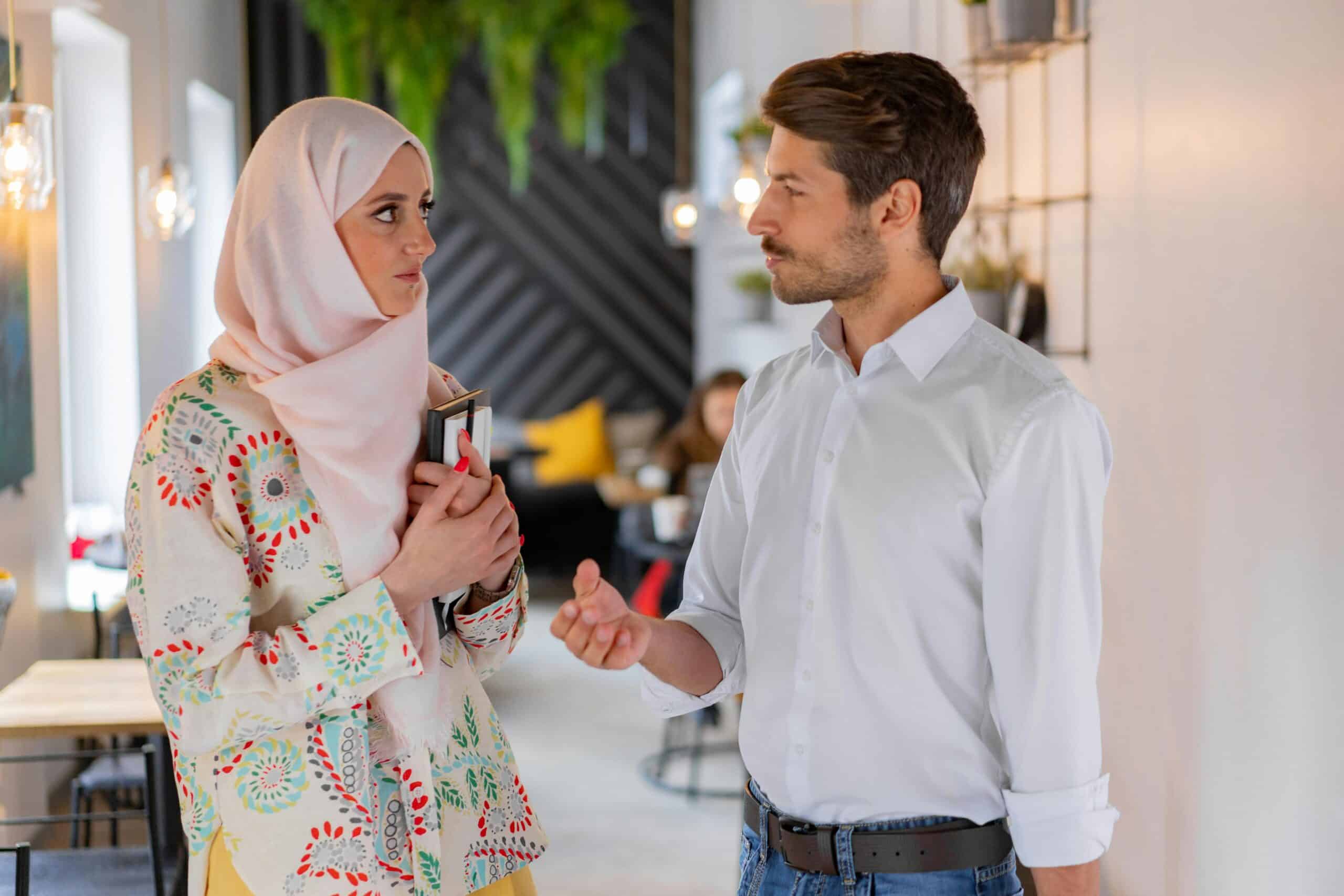 Two colleagues having a conversation in a modern office space, with a woman in a hijab holding notebooks and a man gesturing as he speaks.