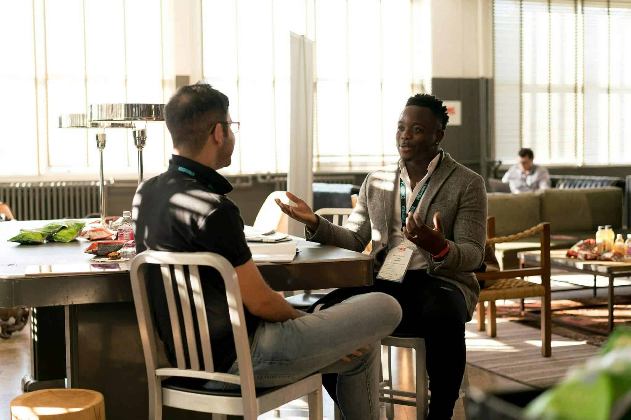 Two men sitting at a table engaged in a conversation, with one man gesturing as he speaks.