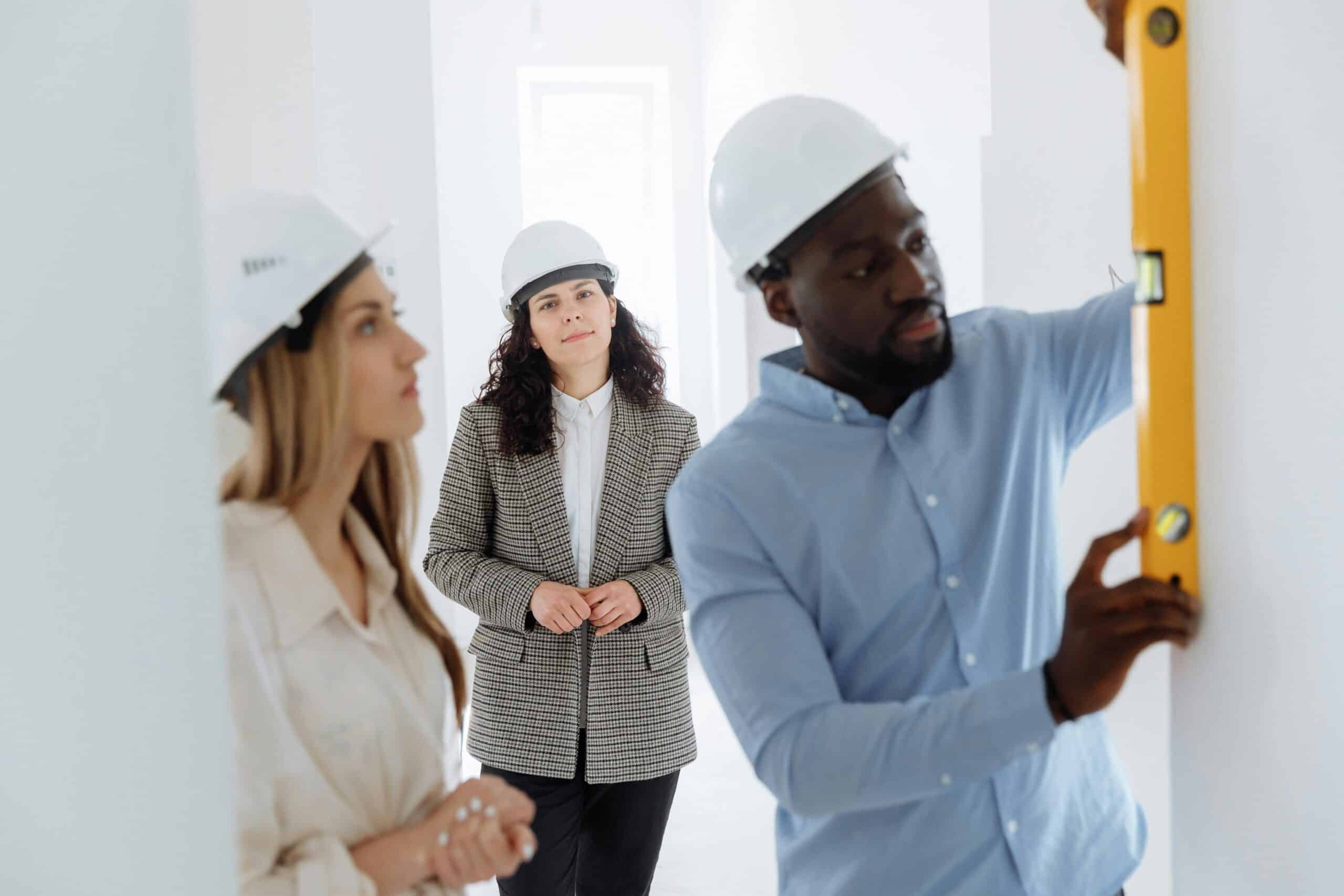 Three people wearing hard hats, including two women and a man, are inspecting a construction site. One woman observes while the man uses a level tool on a wall.