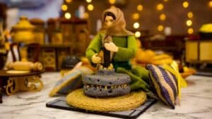 A handcrafted miniature model depicting a traditional scene with a figure in a green outfit and headscarf operating a grinding tool. The setting includes vibrant cushions, a marble surface, and warm lighting in the background.