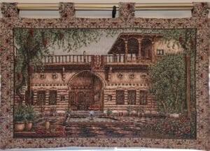 A Gobelin painting featuring a traditional oriental house with intricate architectural details, surrounded by lush greenery and a courtyard with a water fountain. The artwork is framed with an ornate floral border.