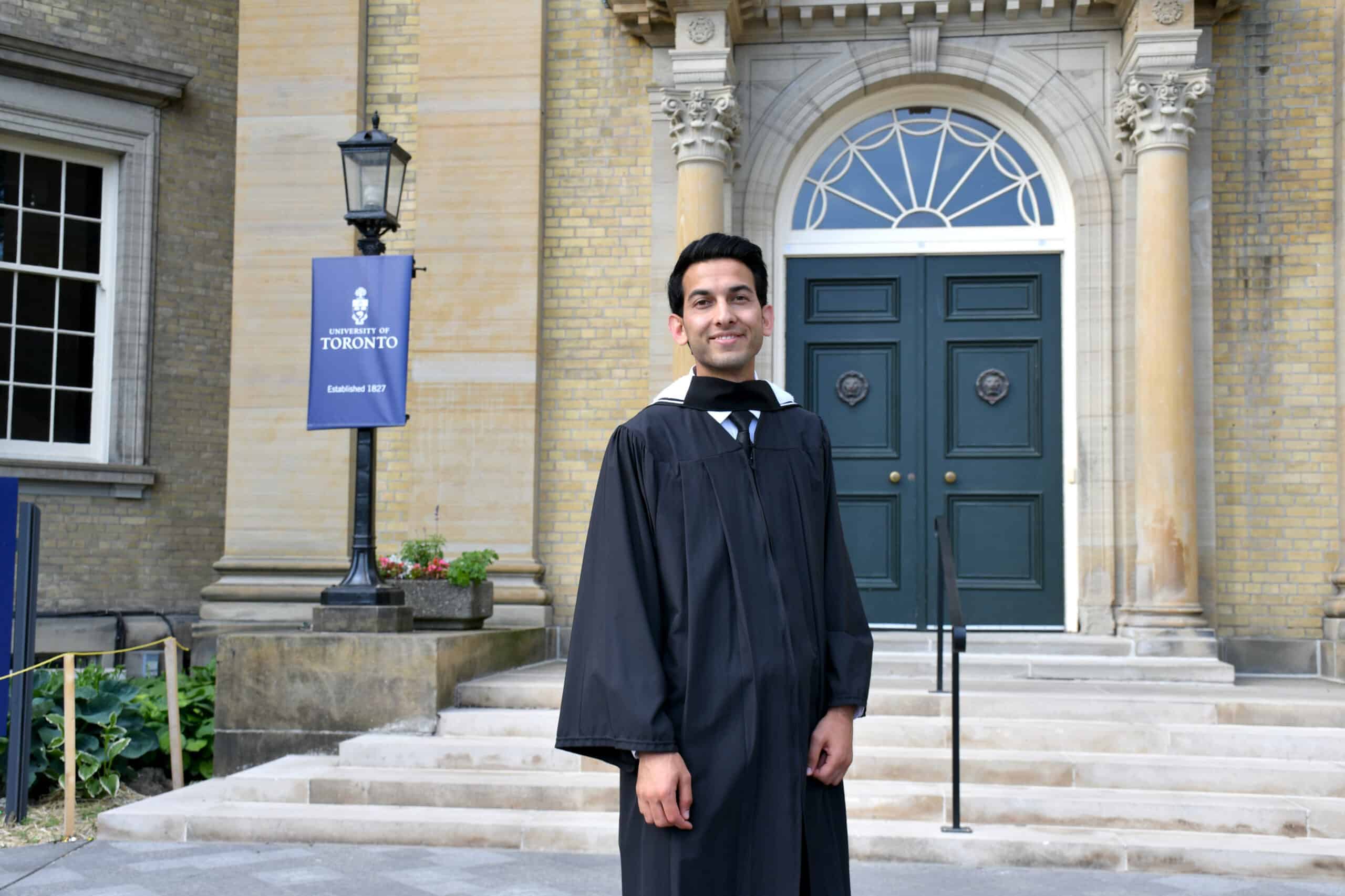 Omer Malikyar in a black graduation gown stands smiling in front of a historic building with columns and double green doors. A University of Toronto banner is visible on the left.