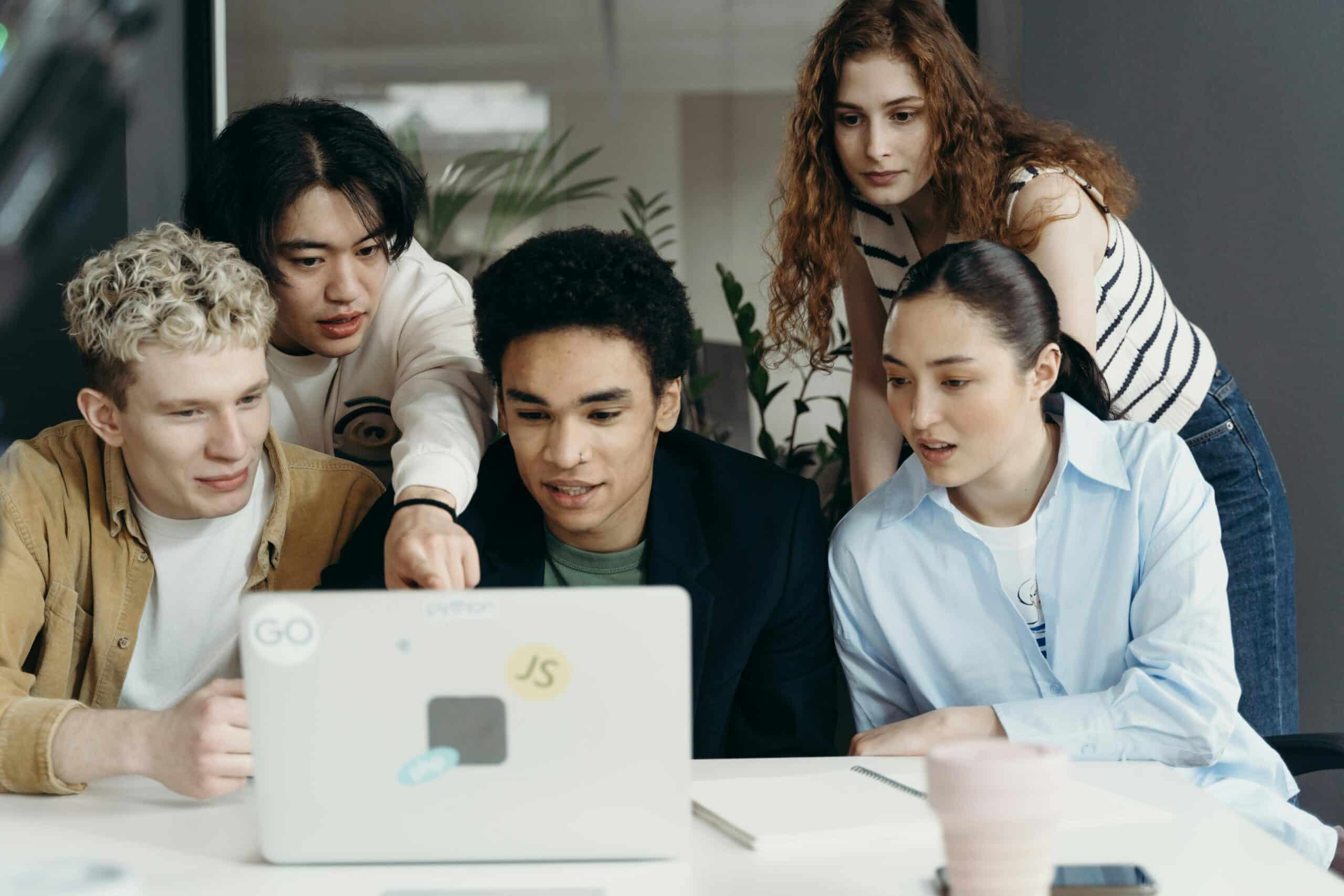 A group of five young adults of diverse backgrounds gathered around a laptop, collaborating and learning together in a bright, modern space