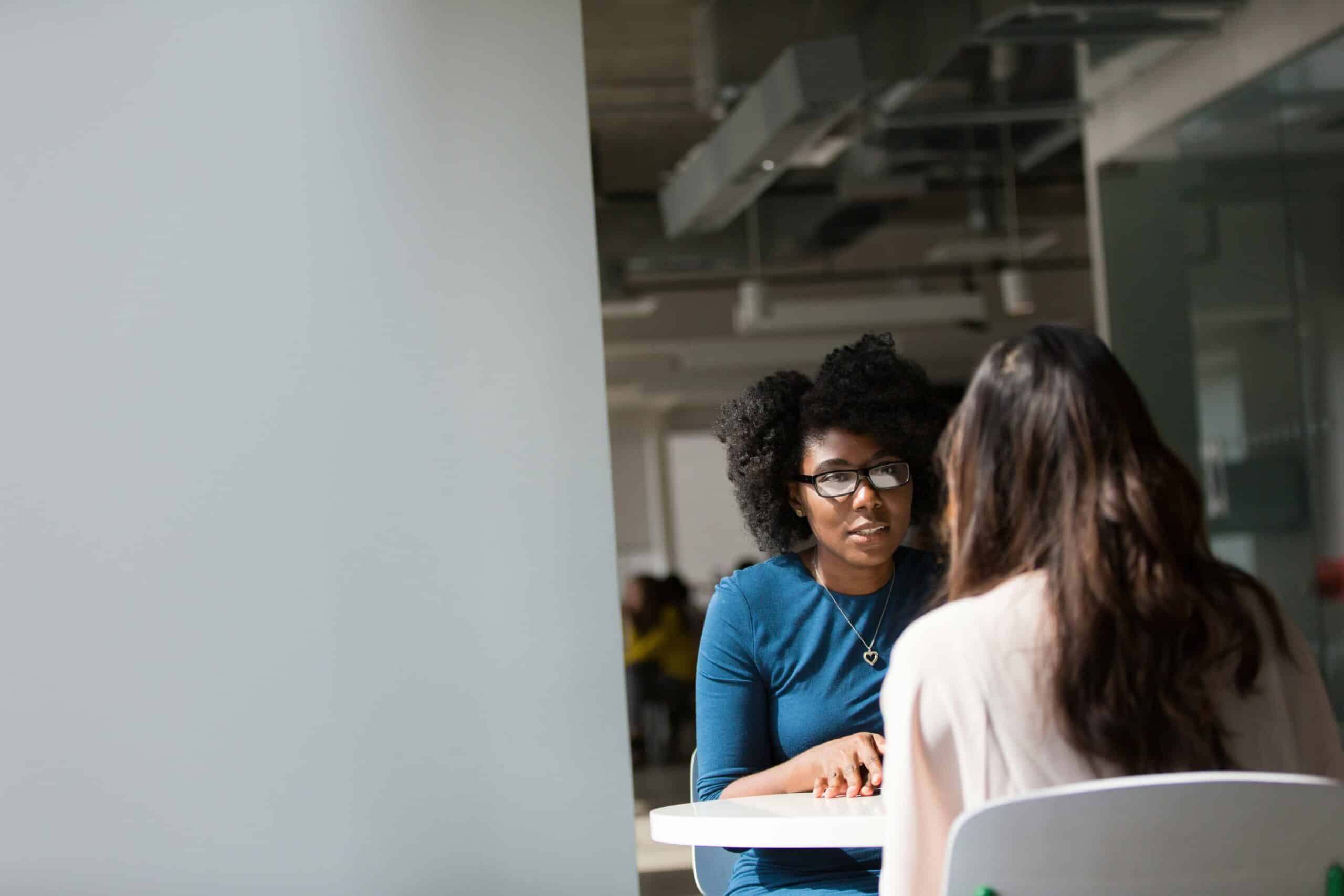 A woman in glasses wearing a blue dress sits across a table from another woman during a mentoring session in a bright, modern office setting.