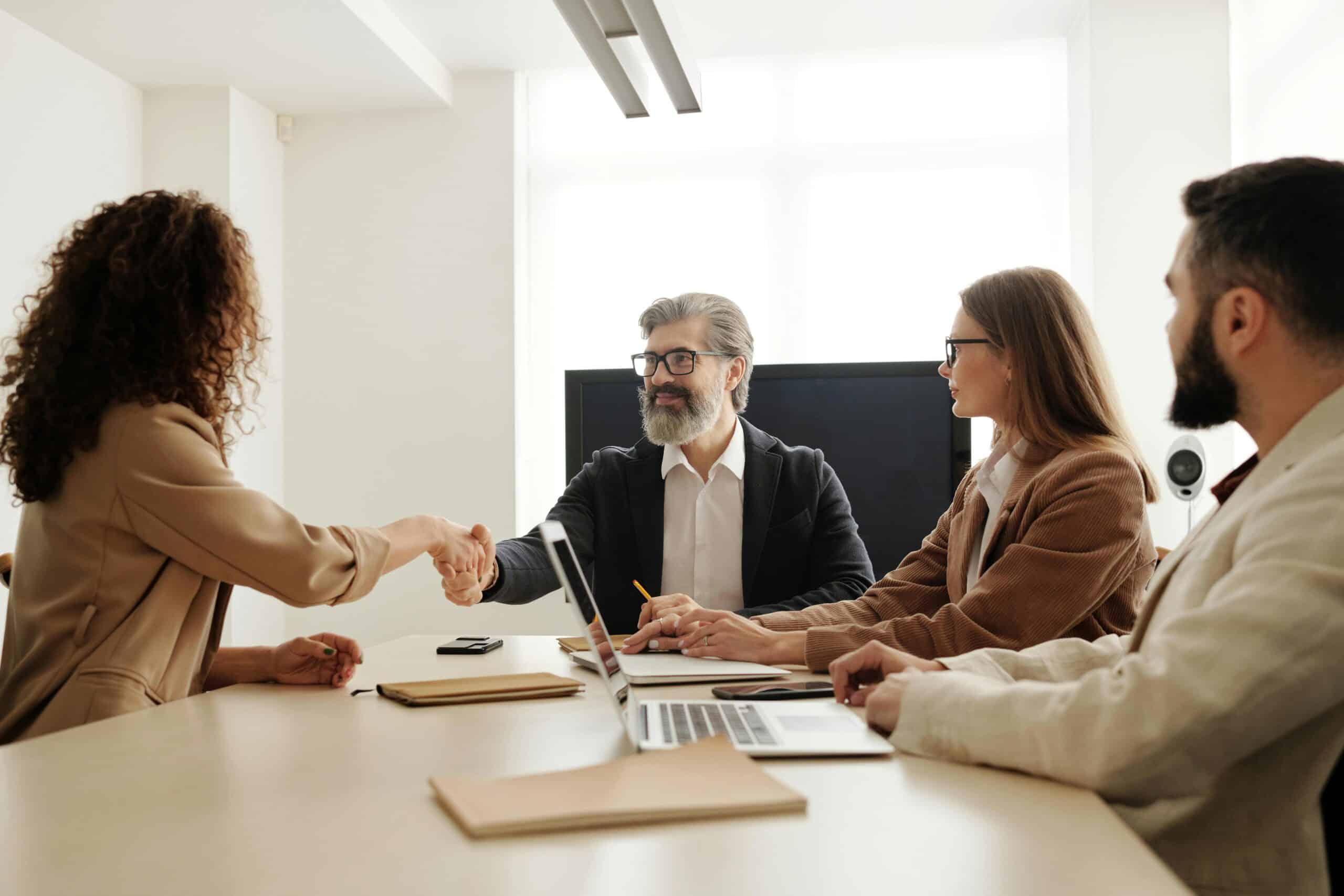 A professional business meeting where a woman in a beige blazer shakes hands with a bearded man in glasses, while two other colleagues observe and take notes.