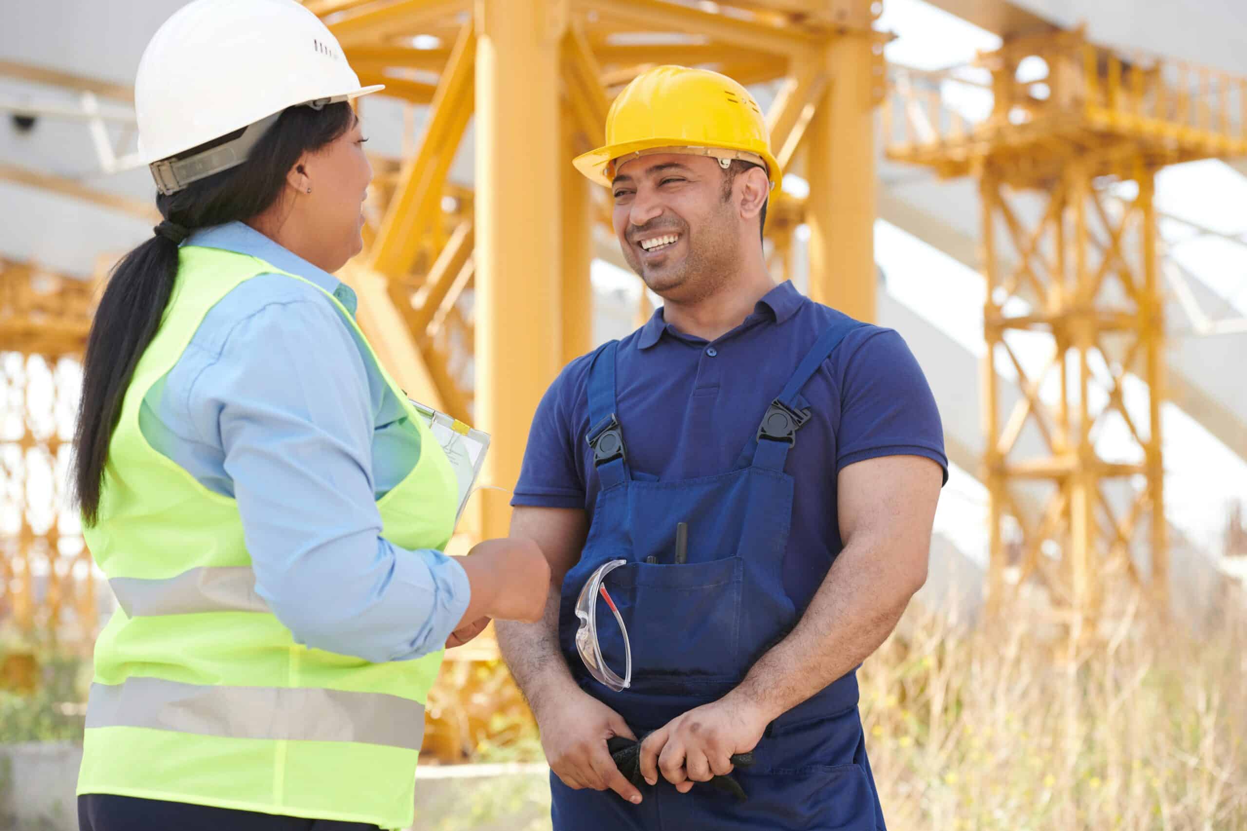 Two construction workers in safety gear smiling and having a conversation at a worksite.