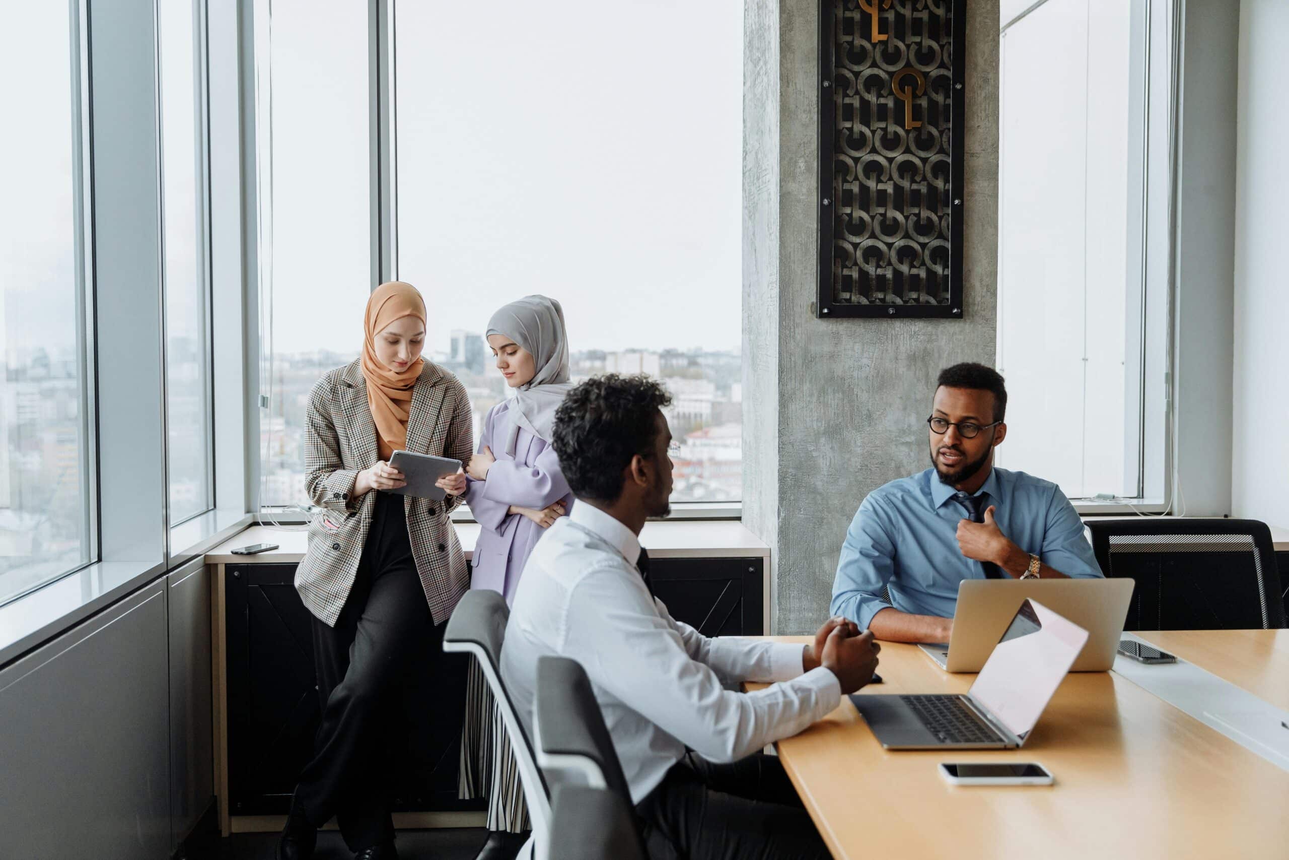 Diverse professionals, including two women wearing hijabs and two men in business attire, engaged in a discussion in a modern office setting with large windows.