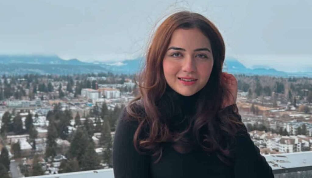 Bhavika Nanda, a young woman with long, wavy brown hair, stands on a high-rise balcony with a wintery cityscape in the background. She wears a black turtleneck sweater and high-waisted denim jeans, smiling warmly at the camera with snow-covered rooftops and mountains visible in the distance.