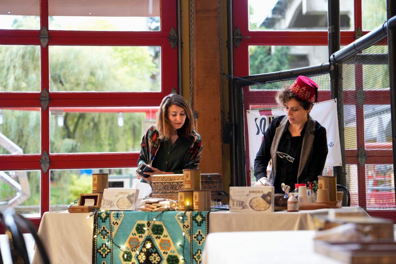 Two women stand behind a market-style display table adorned with a colorful woven textile, offering traditional handmade goods. One woman, wearing a red embroidered hat, is arranging items, while the other, dressed in a dark shawl, interacts with a customer. The table showcases beautifully packaged Middle Eastern sweets, ornate boxes, and artisanal products. A warm, inviting space with red-framed windows and natural light provides a cozy setting.