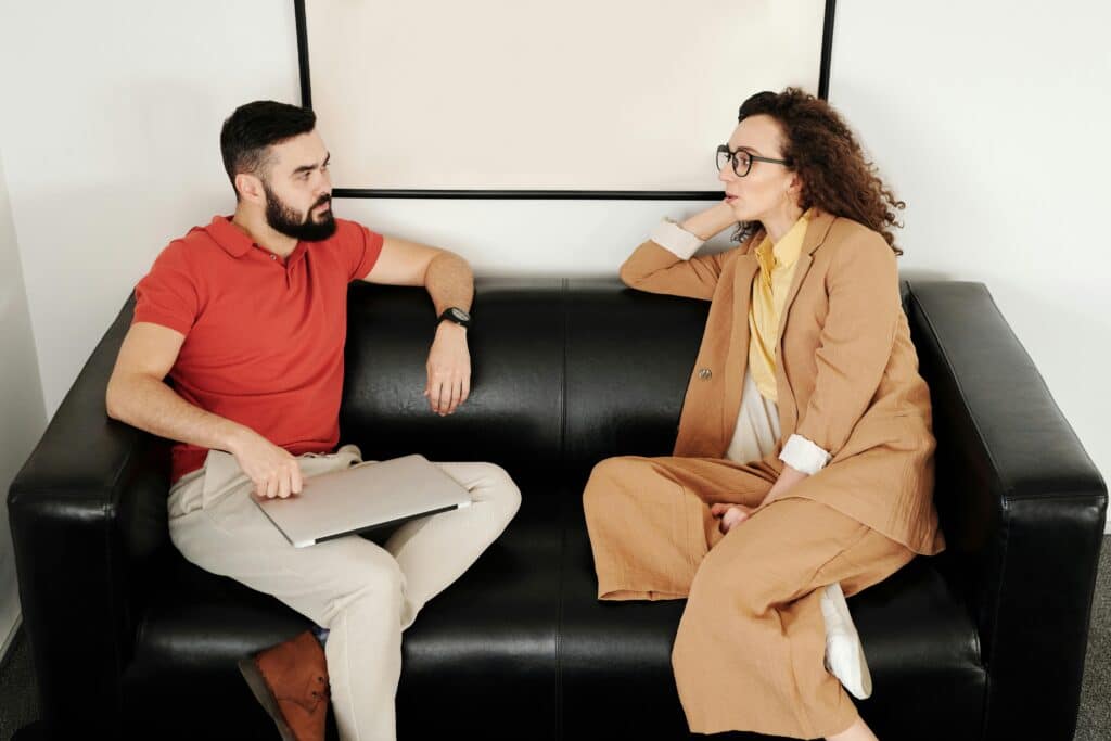 A man and a woman engaged in a professional conversation on a black leather couch, maintaining eye contact and active listening.