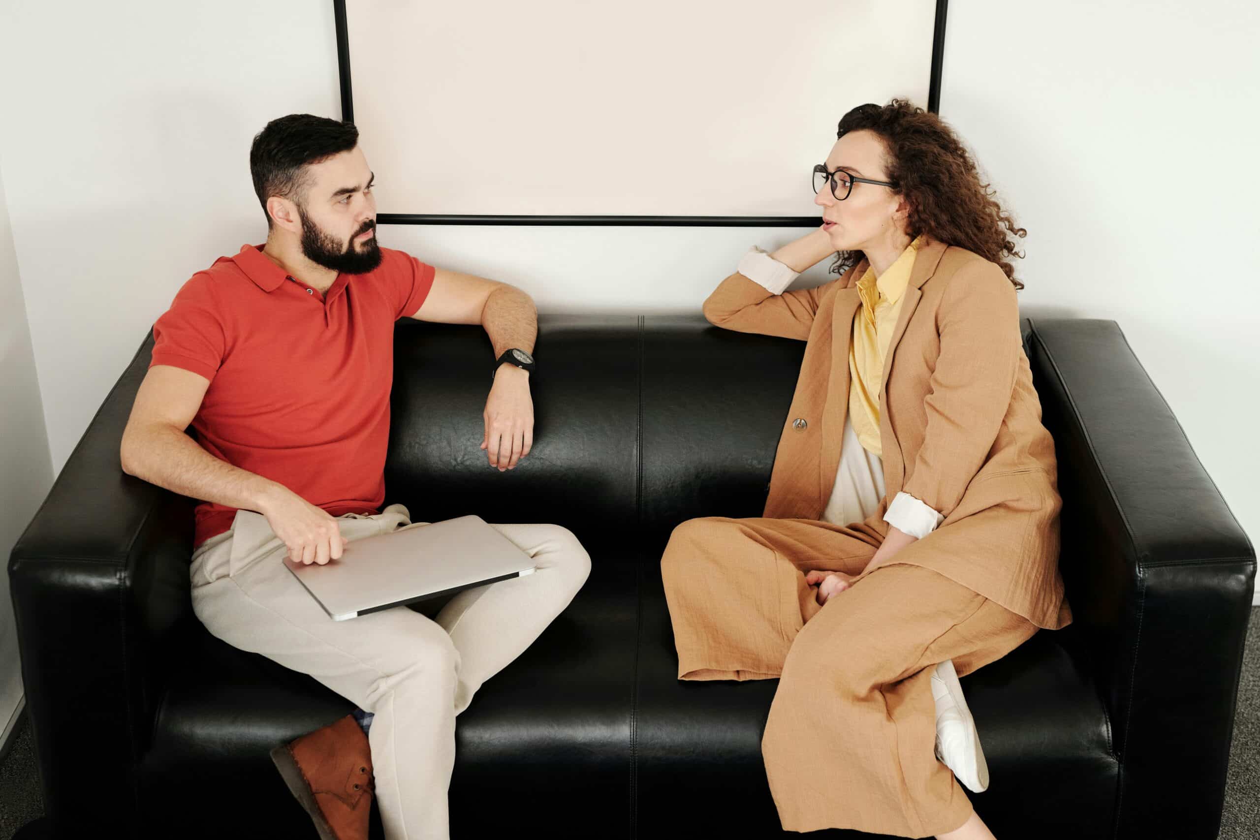 A man and a woman engaged in a professional conversation on a black leather couch, maintaining eye contact and active listening.