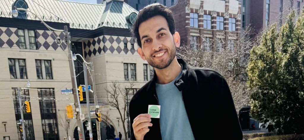 Omer stands proudly outside a polling station holding a voting sticker, smiling with quiet pride after casting his first vote as a Canadian citizen.