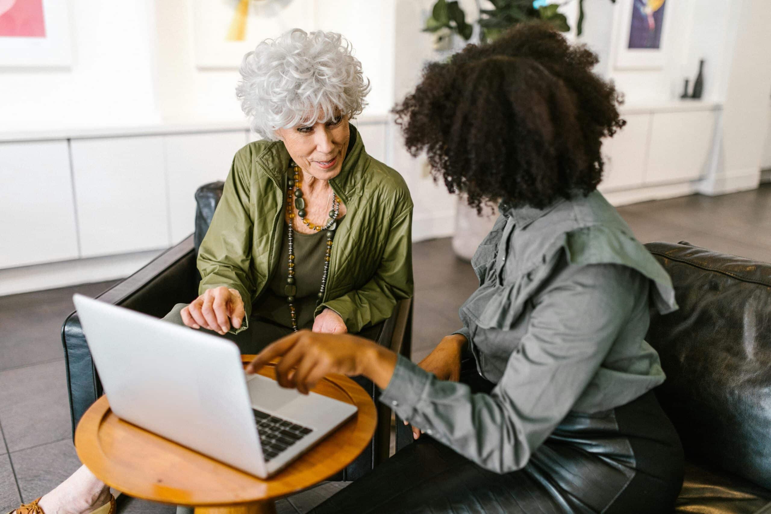 An older woman and a younger woman sit together in a modern lounge, engaged in a collaborative conversation while looking at a laptop screen.