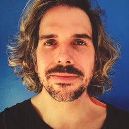 Philip Ackerman, smiling gently with shoulder-length wavy hair and a beard, standing against a bright blue background.