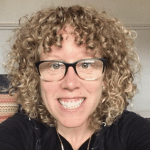 Tanya Aberman, smiling and wearing glasses, with curly blonde hair, seated indoors in front of a neutral background.