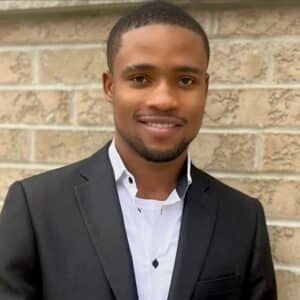 Precious Ezeanyika, a young Black man in a black suit jacket and white shirt, smiling confidently in front of a beige brick wall.