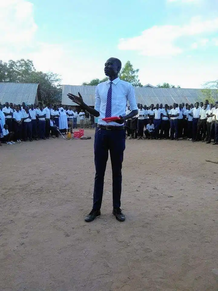 Nhial, a young refugee advocate, stands in front of students at Kakuma Refugee Secondary School in Kenya, passionately addressing the crowd during a school event. Students from diverse backgrounds are seated in an open field, listening attentively.