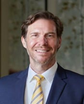 Professional headshot of Shawn M. Bausch, CPA, smiling in a navy suit with a light shirt and yellow striped tie, standing in front of a softly blurred background.