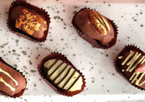 A close-up arrangement of five plump dates that have been dipped in chocolate. The dates are sliced open to reveal their gourmet fillings, which include a green pistachio kunafa mix and a bright orange-flavored filling. The dates are presented neatly on a platter or surface.