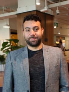 Mustafa Alio, wearing a grey blazer and black shirt, smiling indoors in an office setting.