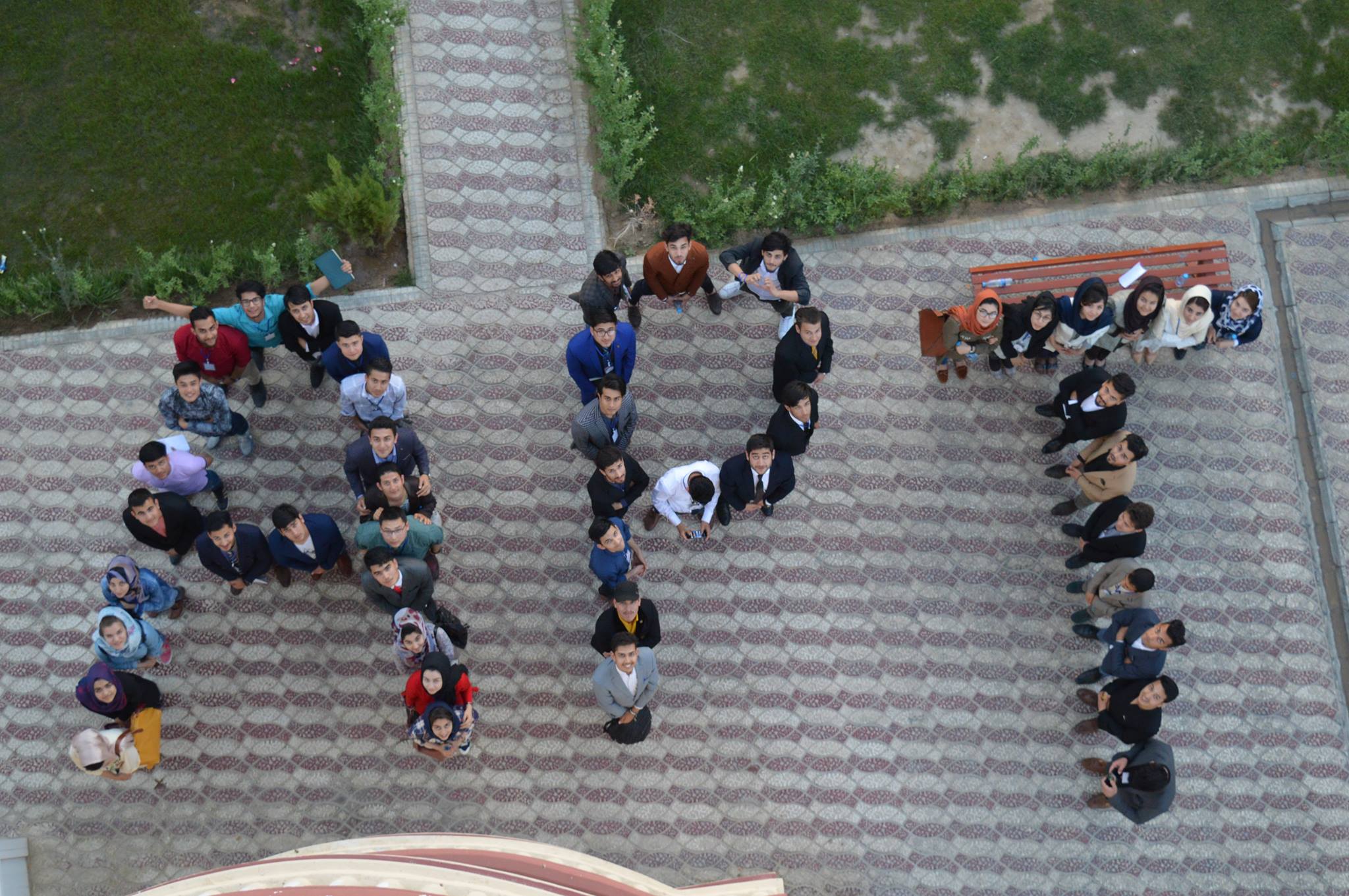 A group of young Afghan men and women standing in formation, viewed from above, forming the initials "APT" on a patterned tile pathway.