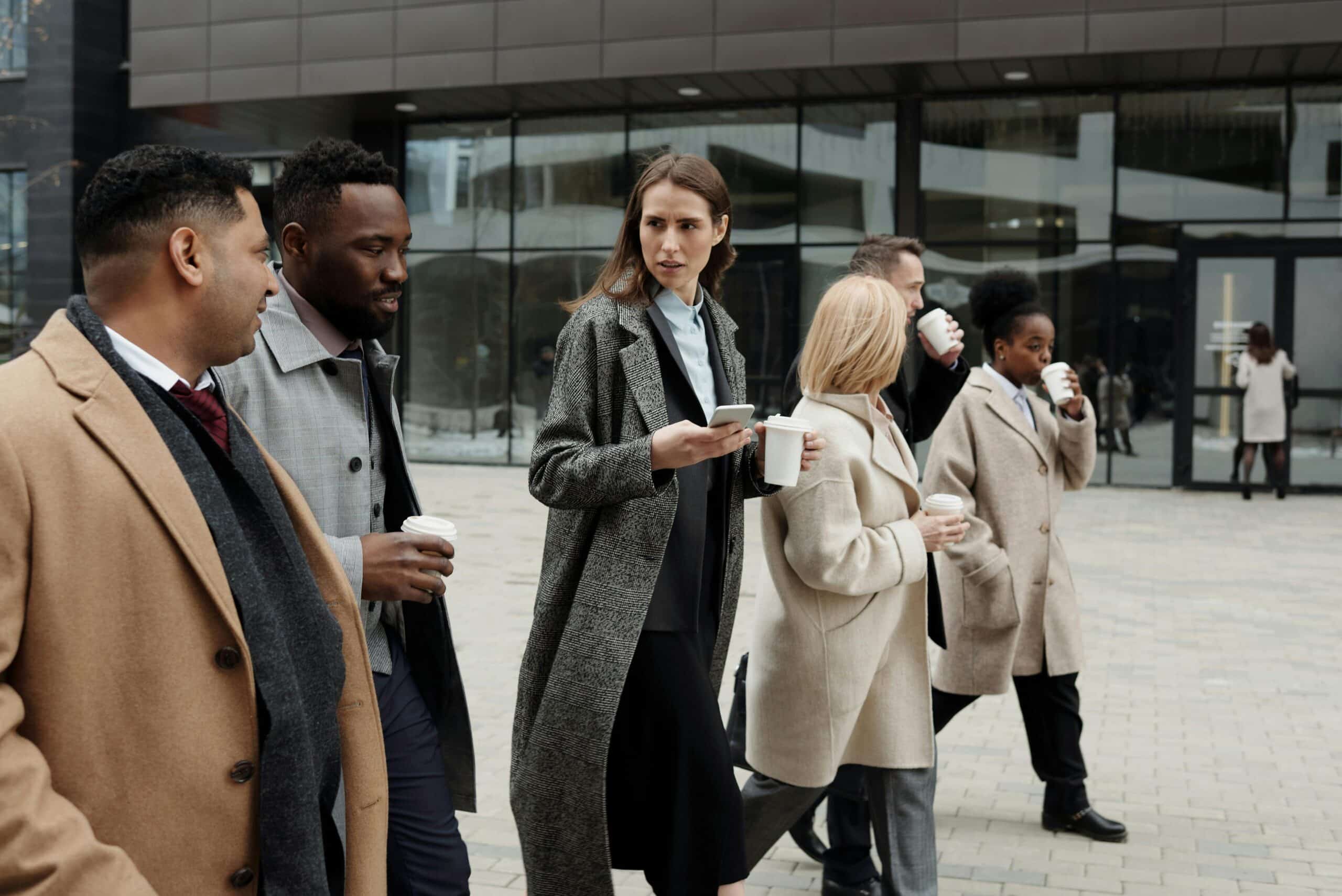 A diverse group of six professionals in formal and business-casual attire walking outdoors while chatting and holding coffee cups. The backdrop features a modern office building with large glass windows.