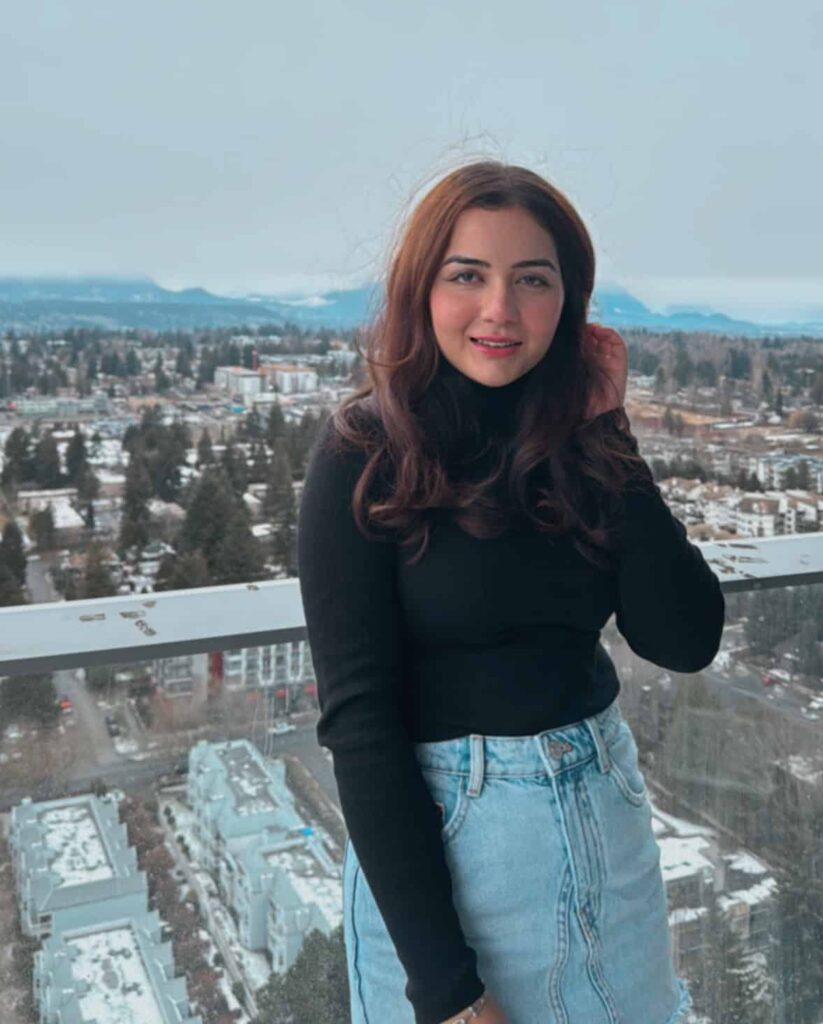 Bhavika Nanda, a young woman with long, wavy brown hair, stands on a high-rise balcony with a wintery cityscape in the background. She wears a black turtleneck sweater and high-waisted denim jeans, smiling warmly at the camera with snow-covered rooftops and mountains visible in the distance.