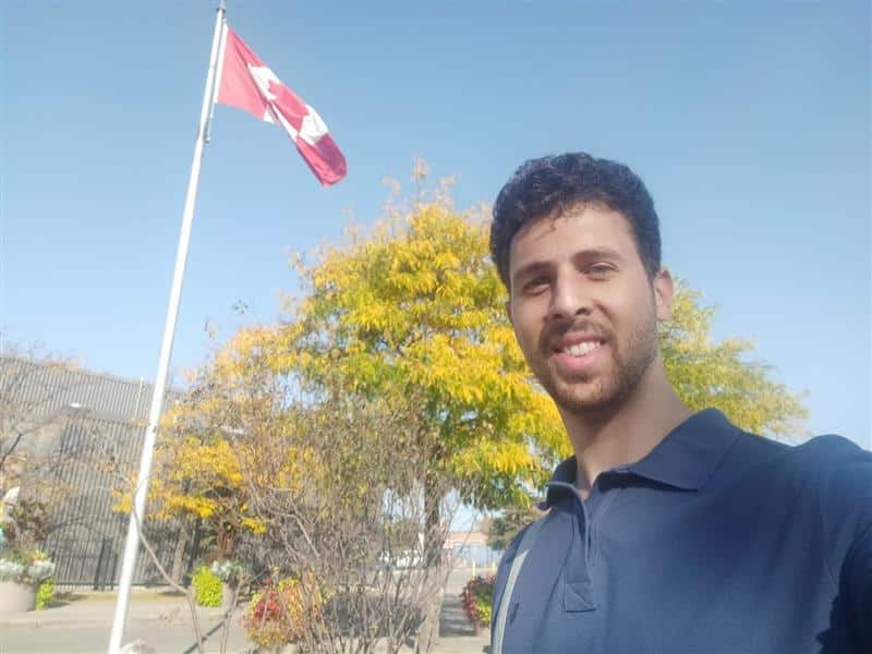 A smiling young man stands outdoors in front of a Canadian flag and autumn trees, capturing a moment of pride and belonging in his new home.